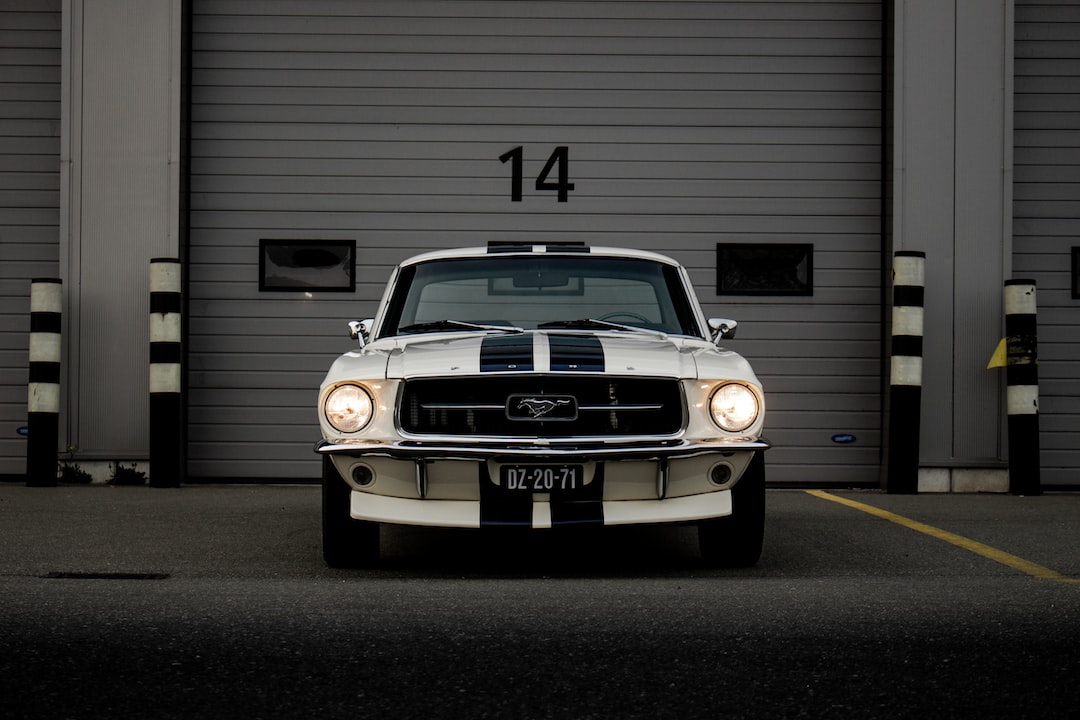 Un Ford Mustang clásico blanco con rayas azules de competición, que encarna el lujo, está aparcado frente a la puerta gris de un garaje marcada con el número 14.