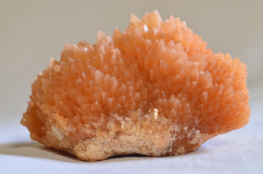Un racimo de cristal rosa anaranjado con puntas afiladas y triangulares se asienta sobre una superficie clara sobre un fondo neutro, irradiando un aire de lujo y elegancia.